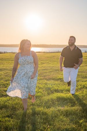A couple walks through a sunlit field at golden hour near the waterfront.