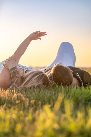 A couple lies in grass during sunset with arms raised against golden sky.