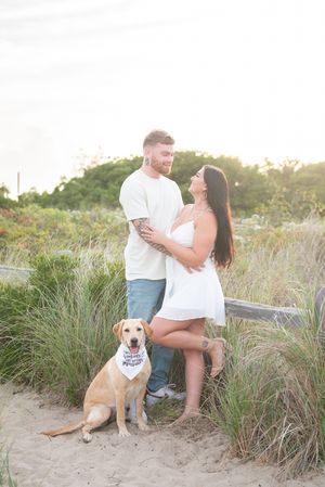 A couple poses with their golden retriever wearing a bandana on a sandy beach path.