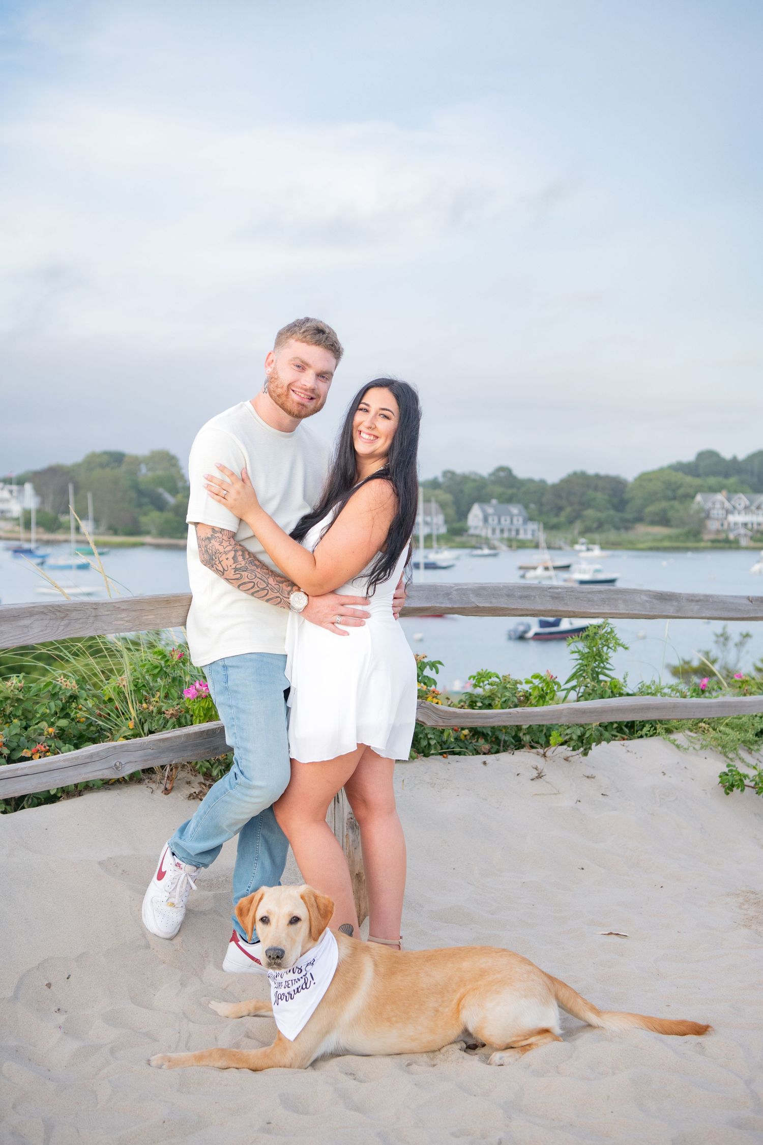 A couple poses with their dog by a harbor with boats and coastal homes in the background.