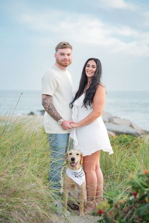 A couple stands with their golden retriever on a grassy coastal path overlooking the ocean.