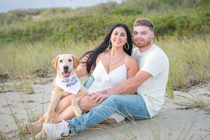 A couple sits casually with their golden retriever on a sandy beach path surrounded by dunes.