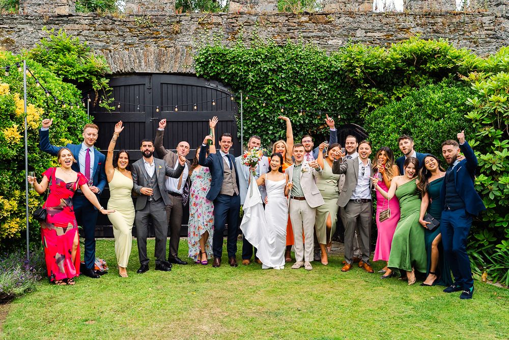 Large wedding party group photo posing together in front of stone wall and garden.