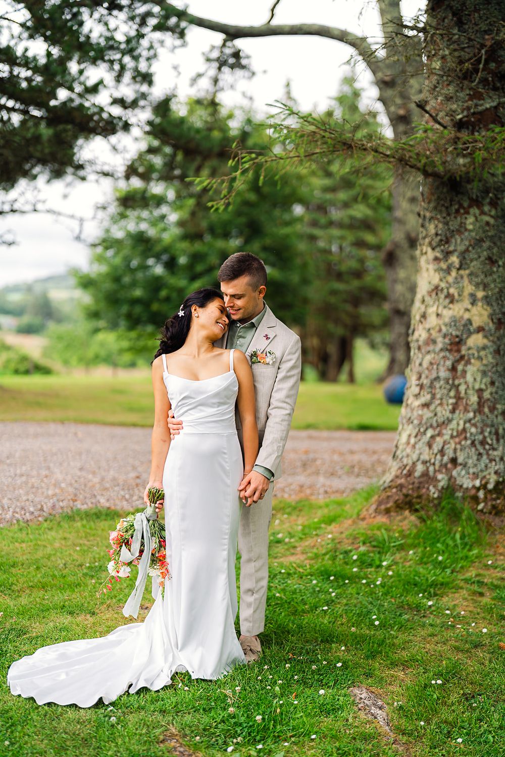 Romantic wedding portrait of couple standing under large tree in elegant attire.