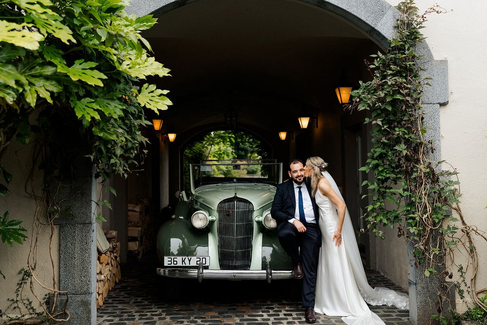 Classic wedding portrait featuring vintage car under stone archway with hanging lanterns.