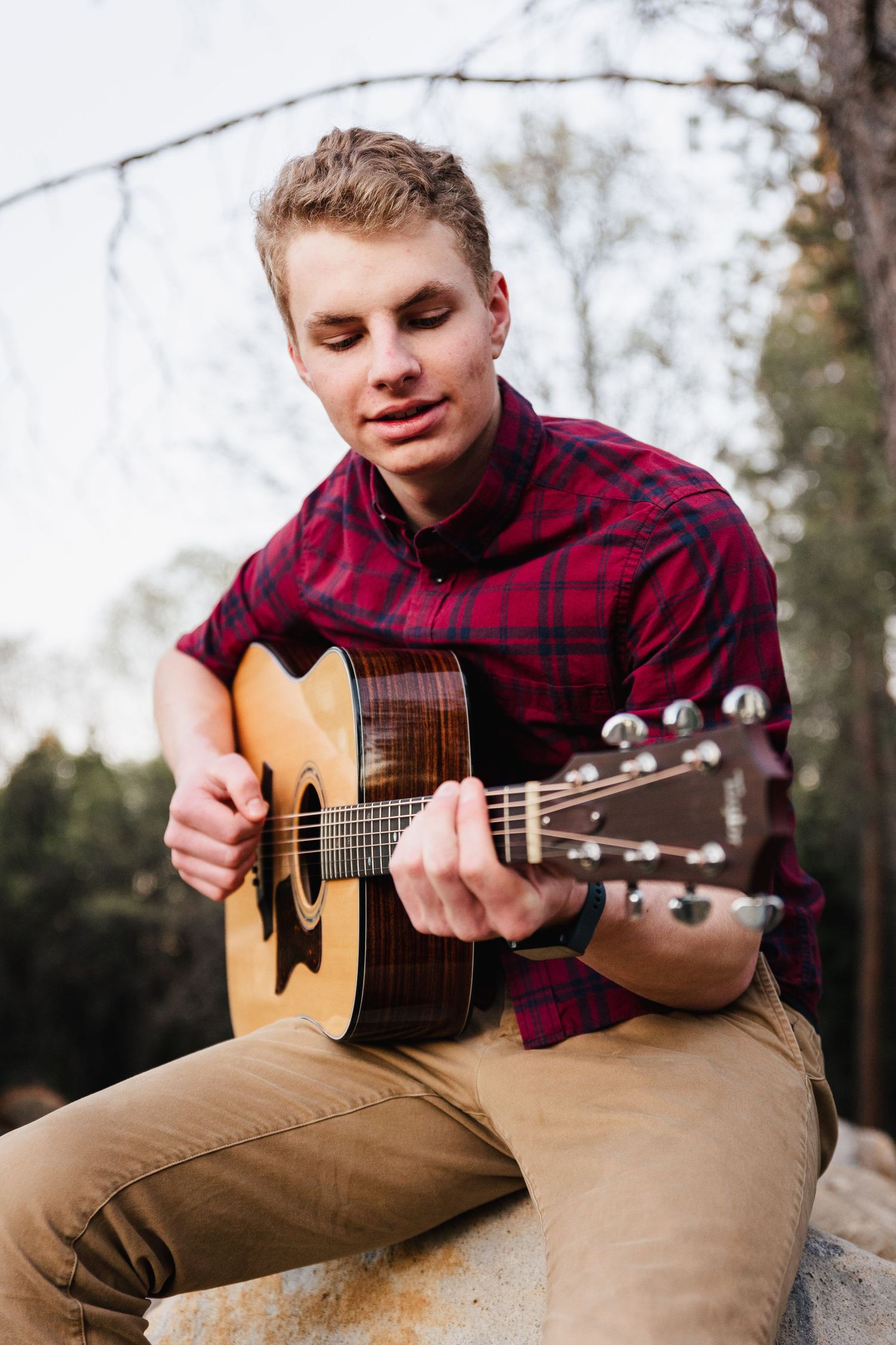A musician in a plaid shirt plays acoustic guitar while sitting outdoors in grass valley, California.