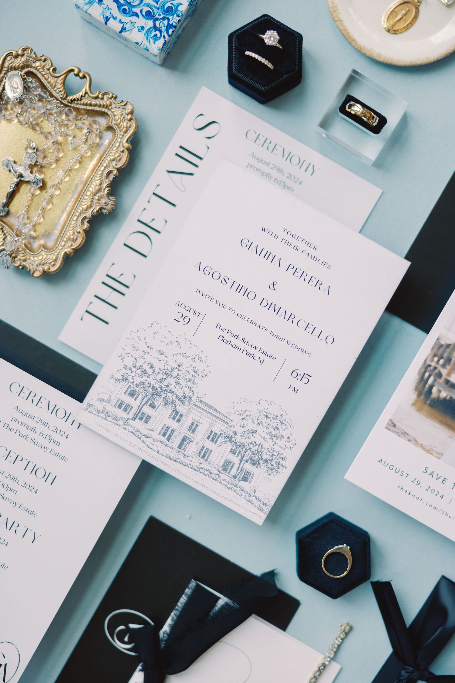 Elegant wedding stationery flatlay with navy and white invitation suite, diamond rings, and accessories on a blue background.