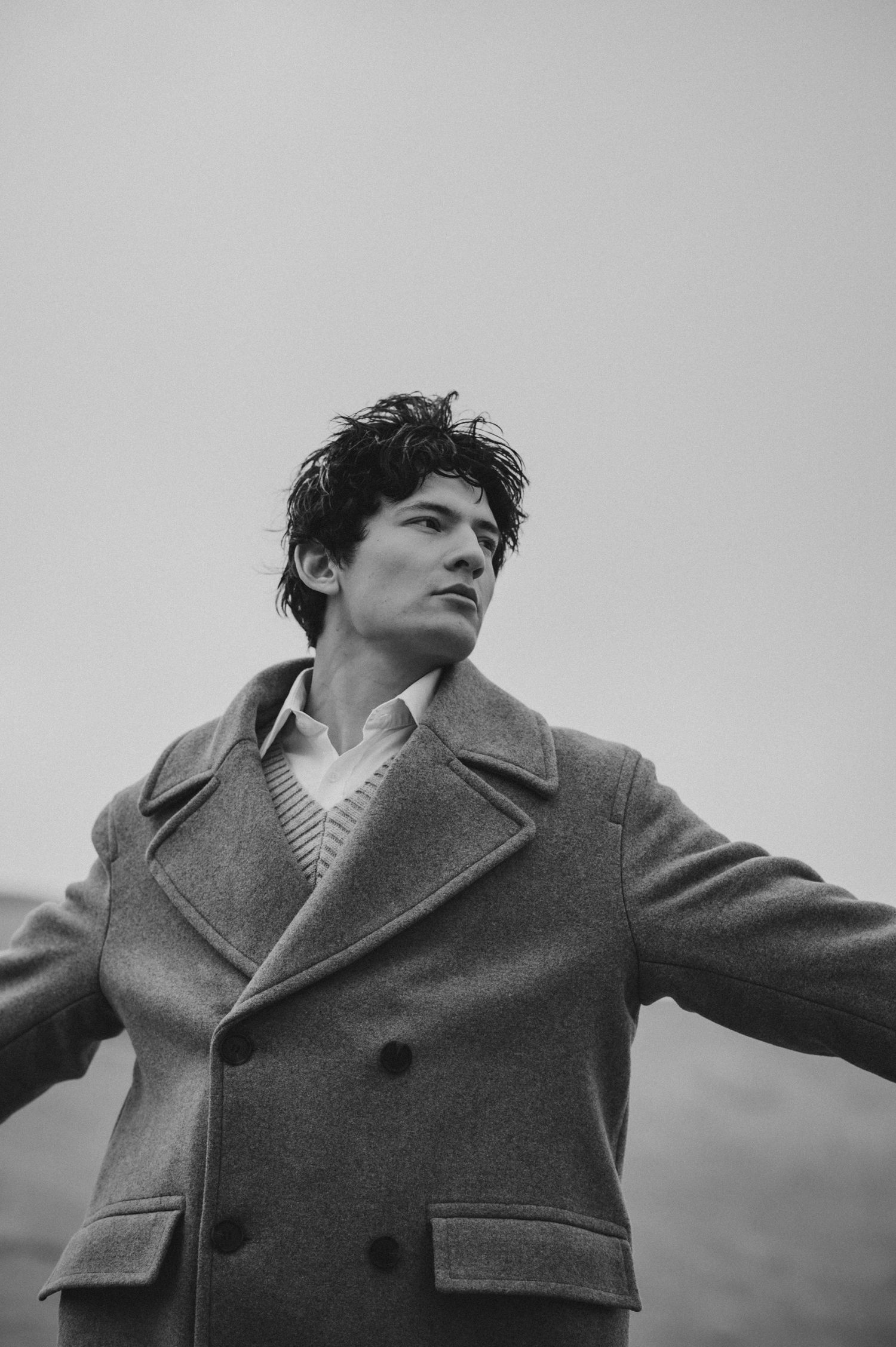 A black and white portrait of a person wearing a wool overcoat with arms outstretched against an overcast sky.