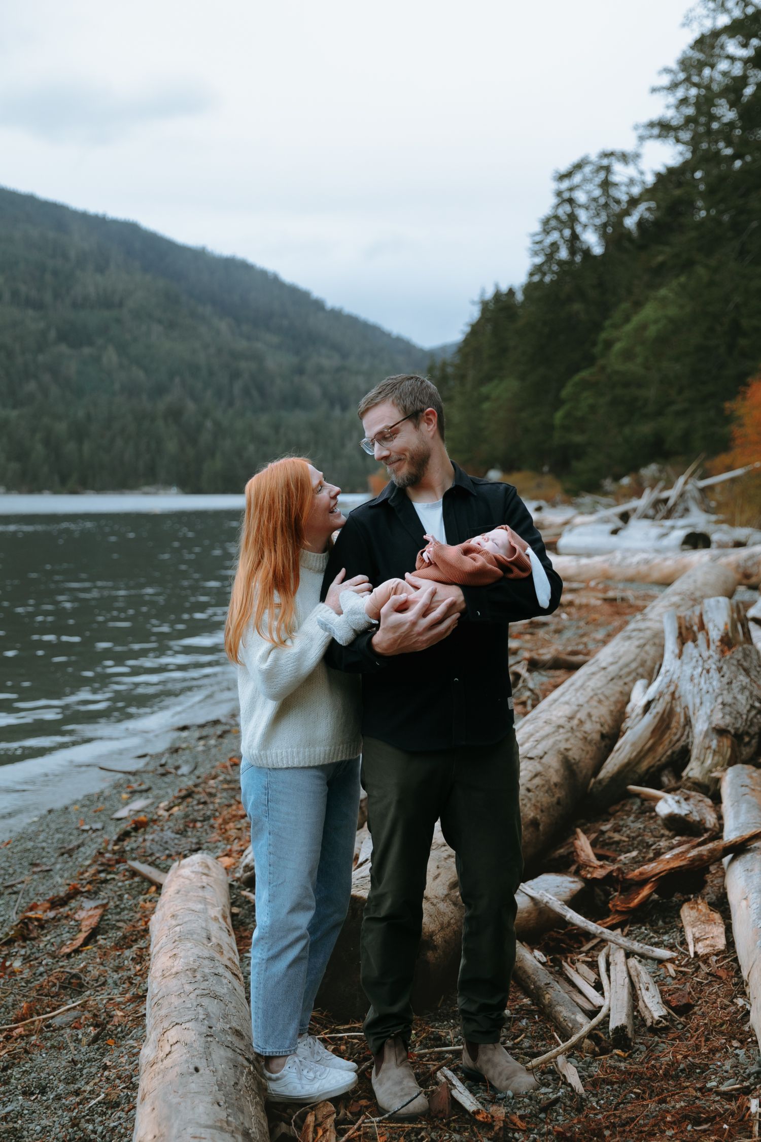 A couple stands on a rocky lakeshore with mountains and evergreen forest in the background on an overcast day.