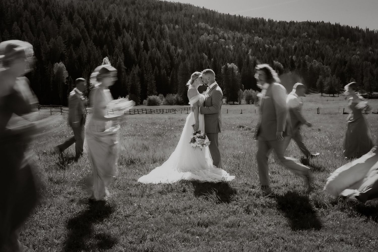 Black and white photo of a wedding celebration with people dancing together in a circle on a grassy field with mountains behind.