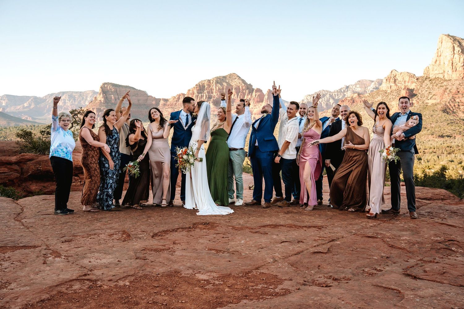 Wedding party celebrates on red rock formation in Sedona, Arizona, with stunning desert landscape and mountains in background.