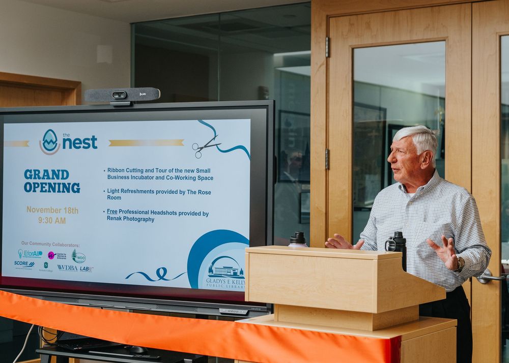 A speaker presents at a grand opening event with a Nest company logo displayed on a screen beside an orange table.