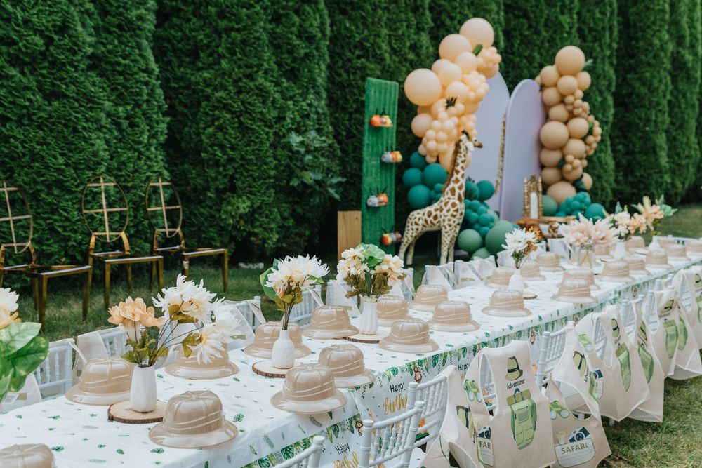 Elegant garden party table setting with gold balloons, white linens, and floral centerpieces against a lush green hedge wall.