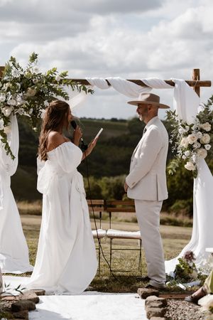 Wedding guests dressed in all white gather outdoors under a wooden arch decorated with white fabric and flowers.