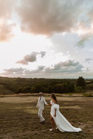 A couple in white wedding attire walks across a scenic field at sunset with dramatic clouds in the background.