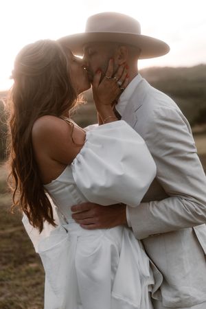 A romantic couple in white outfits share a tender kiss at sunset, with the person wearing a wide-brimmed hat.