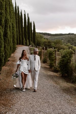 A couple walks down a cypress tree-lined gravel path during a romantic outdoor wedding photoshoot.