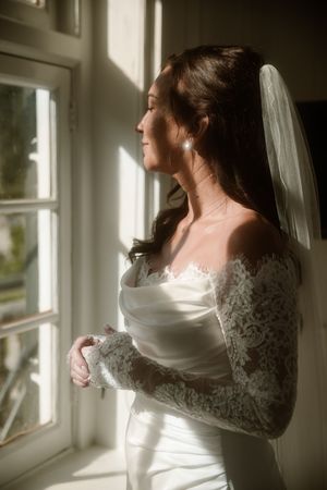 A bride in a long-sleeved lace wedding dress stands by a sunlit window in a series of romantic poses.