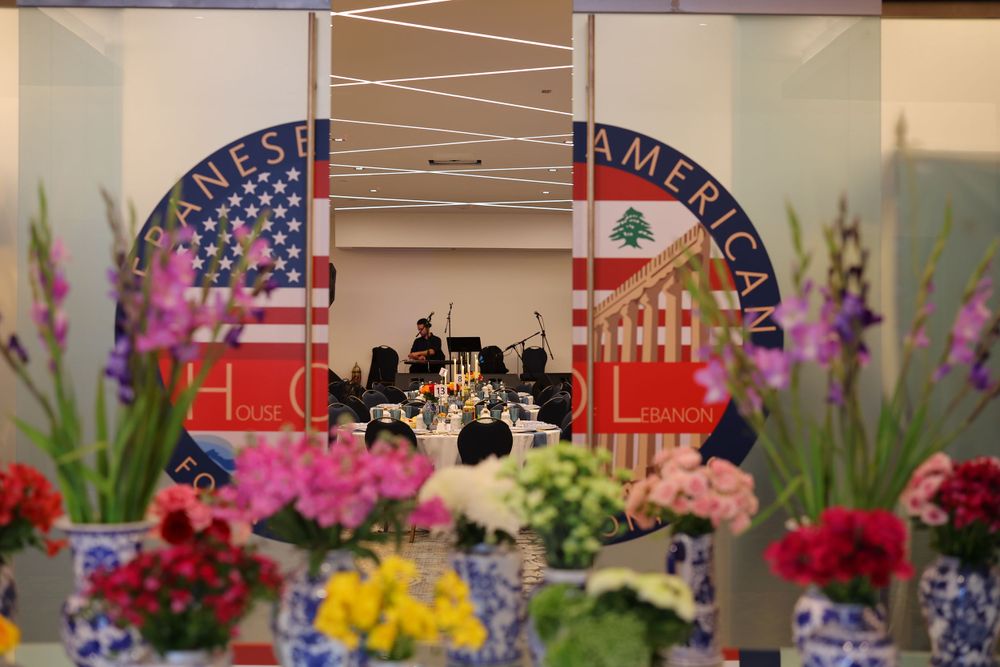 American and Lebanese flags frame a stage with floral arrangements in blue and white vases.