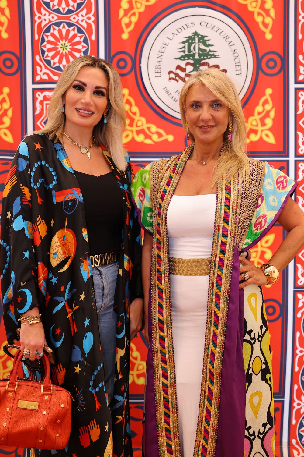 Two women pose in colorful outfits against a decorative red and orange patterned wall with Arabic calligraphy.