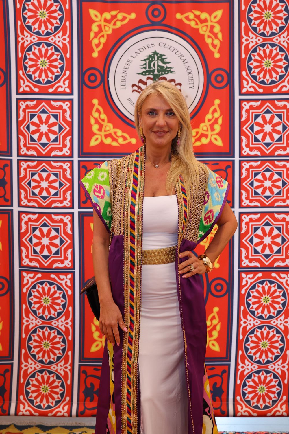Woman in white dress and colorful kimono-style robe poses against vibrant Middle Eastern wall design.