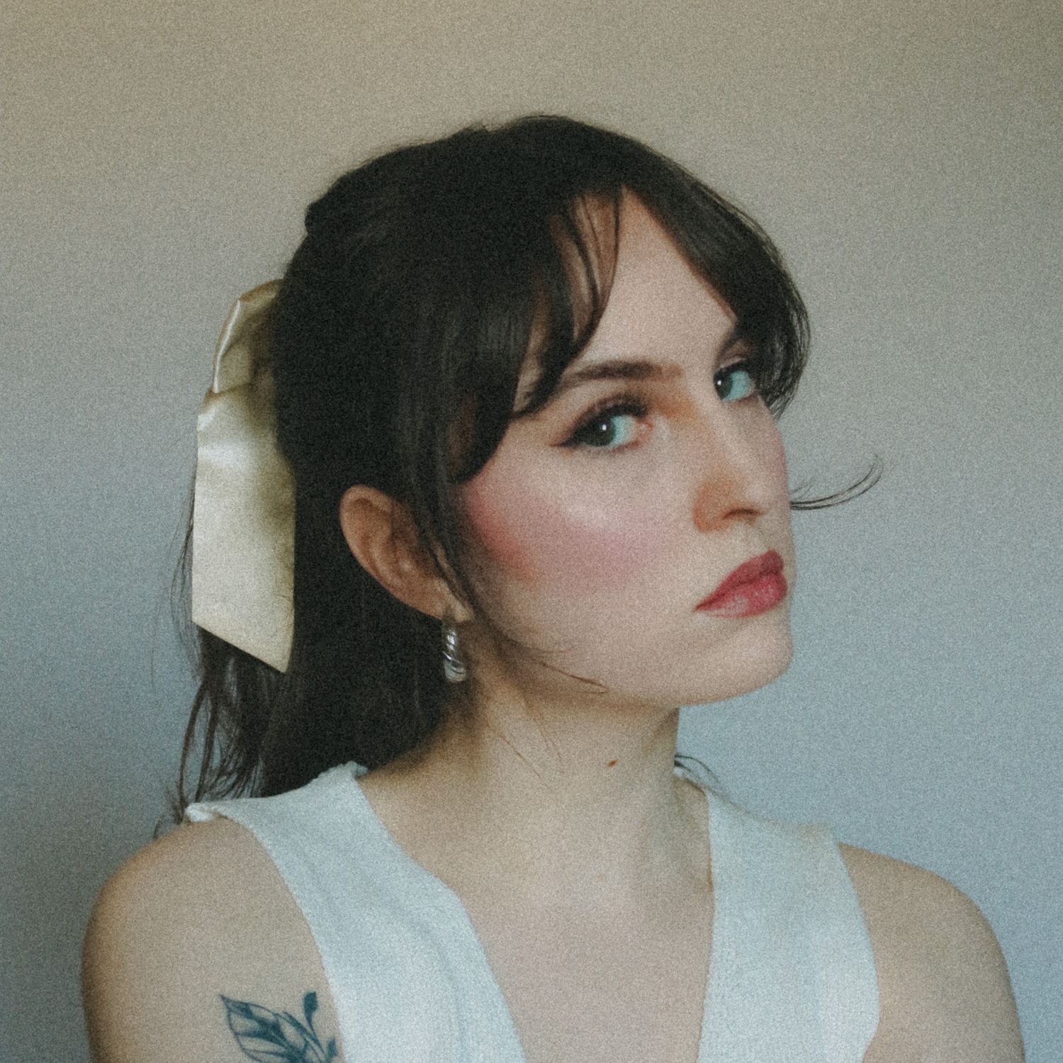 Portrait of someone with dark hair, red lipstick and a white bow hair accessory against a gray background.