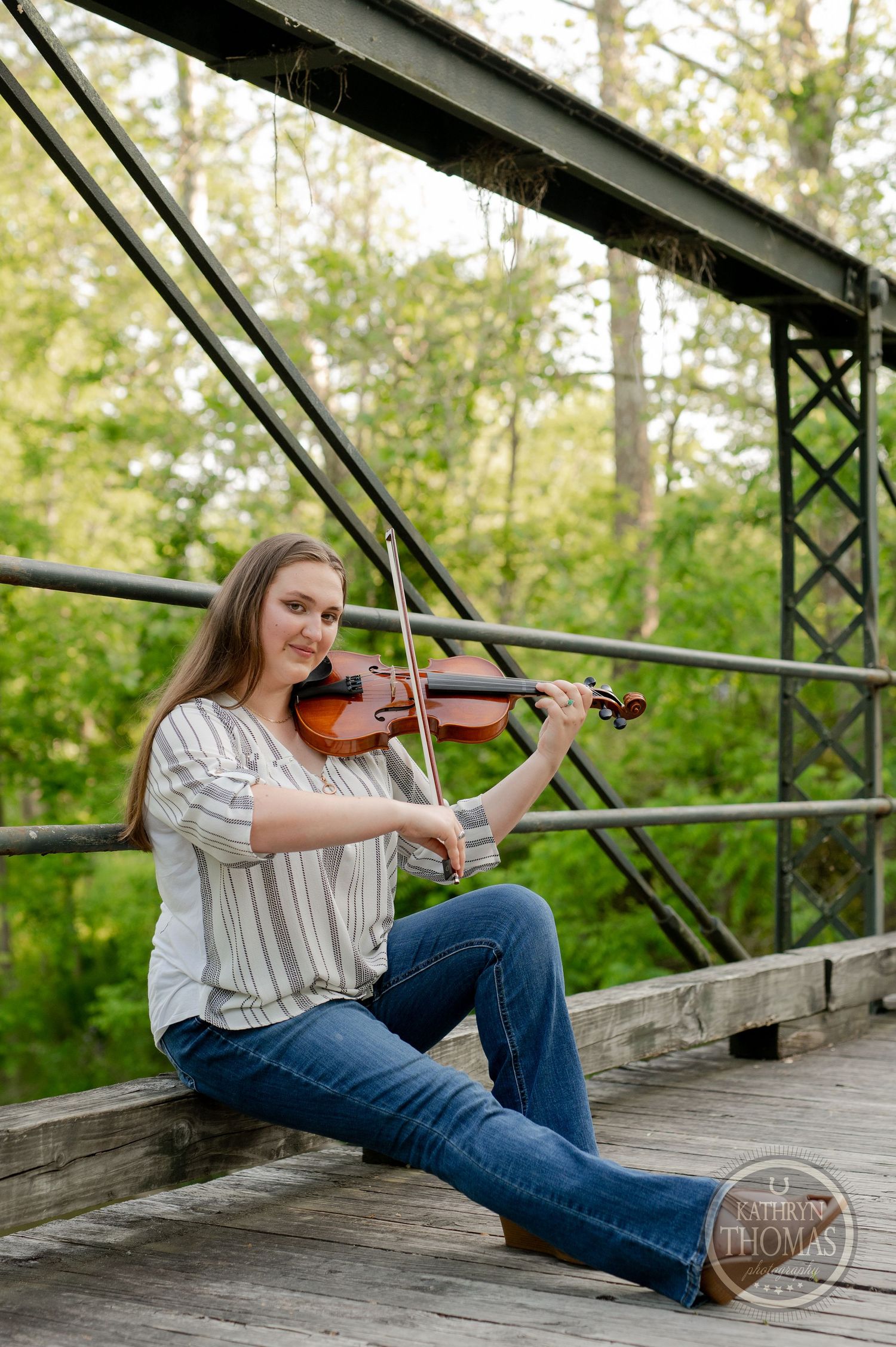 Musician plays violin while seated on wooden bridge with metal railings.
