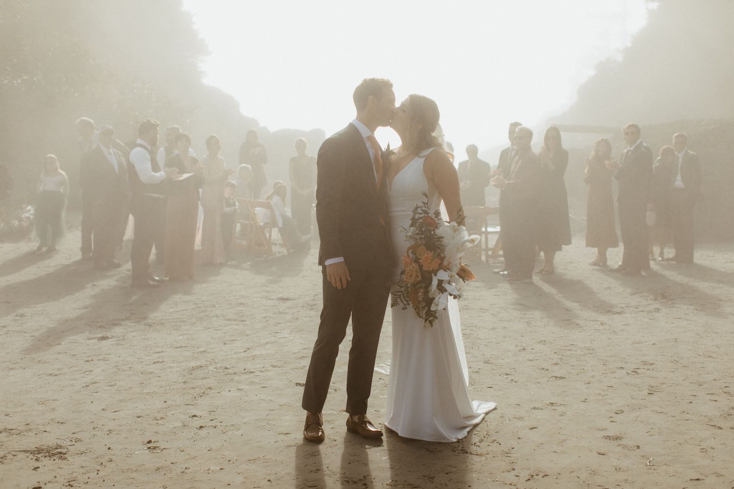 Newlyweds share first kiss in misty beach setting surrounded by wedding guests at sunset.