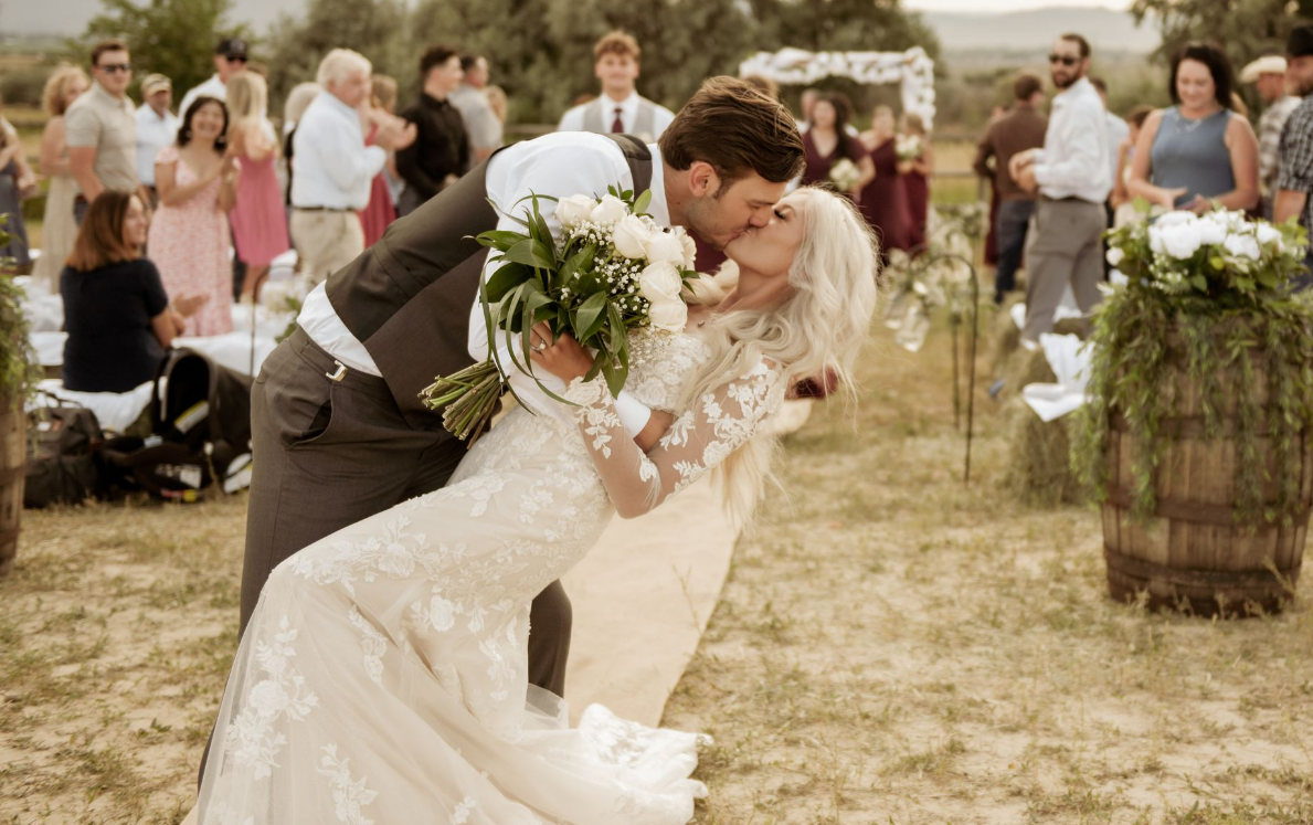 Romantic wedding scene with couple sharing a dip kiss outdoors among guests.