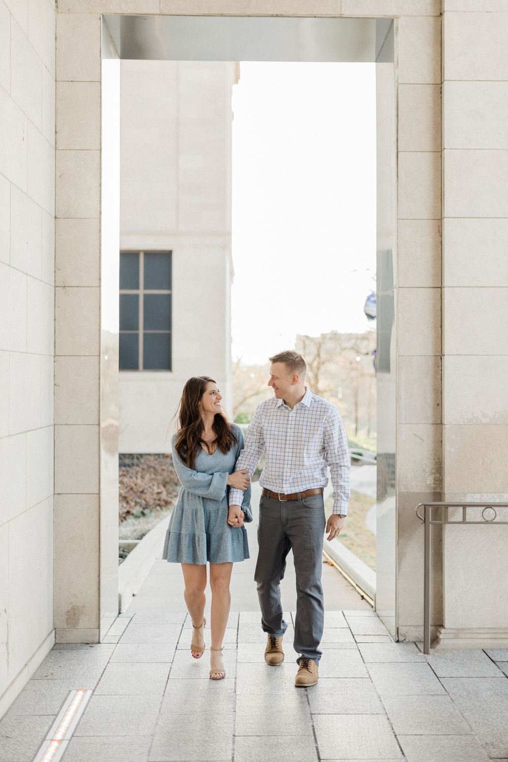 A couple walks through a columned archway at a museum during their engagement shoot.