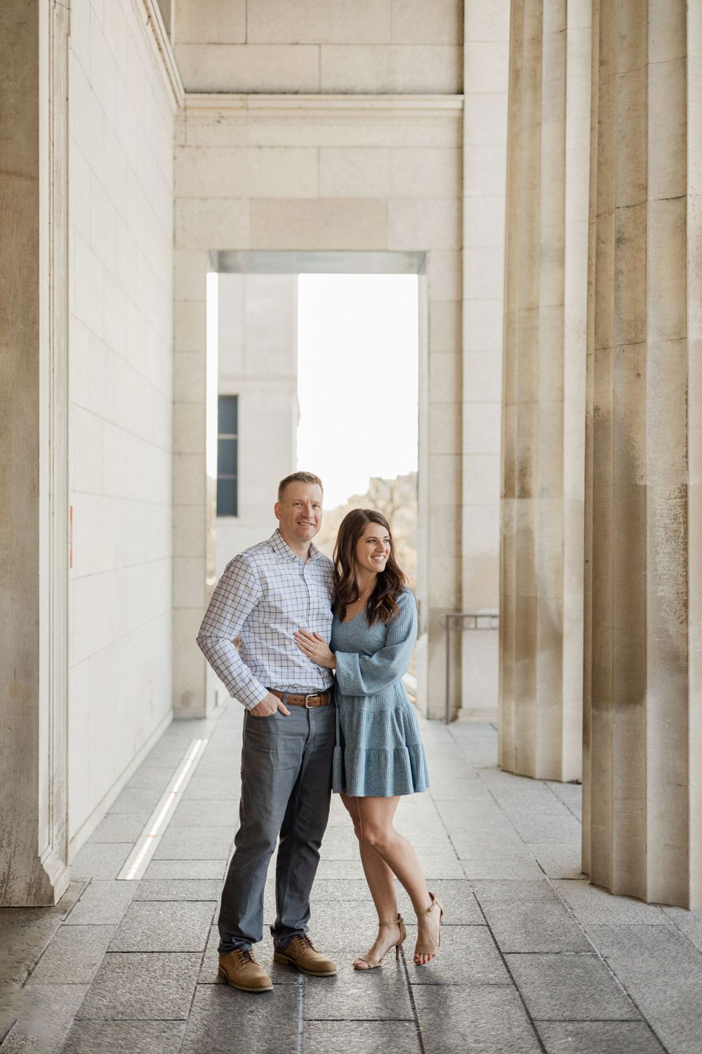 A couple poses between grand stone columns during an engagement photo session.