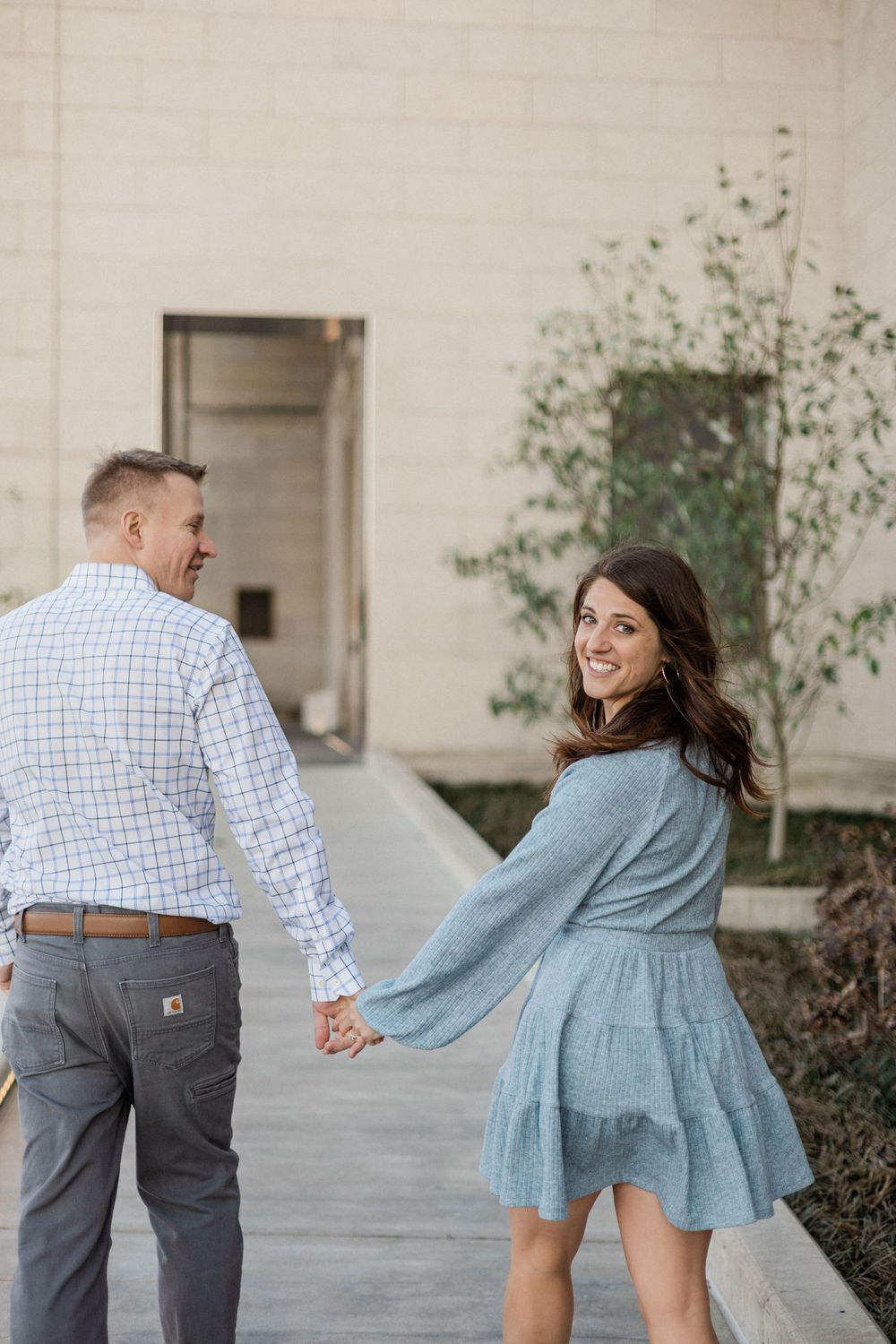 Playful couple walking and holding hands along a concrete walkway next to a stone building.