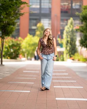 A person in a floral top and wide-leg jeans walks along a brick pathway surrounded by greenery and buildings.
