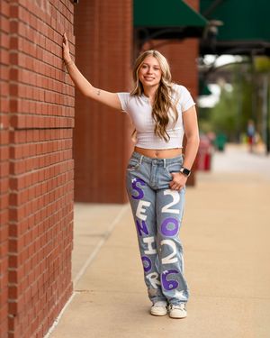 Person wearing ripped jeans with purple and white graphics leans against brick wall in casual outdoor setting.
