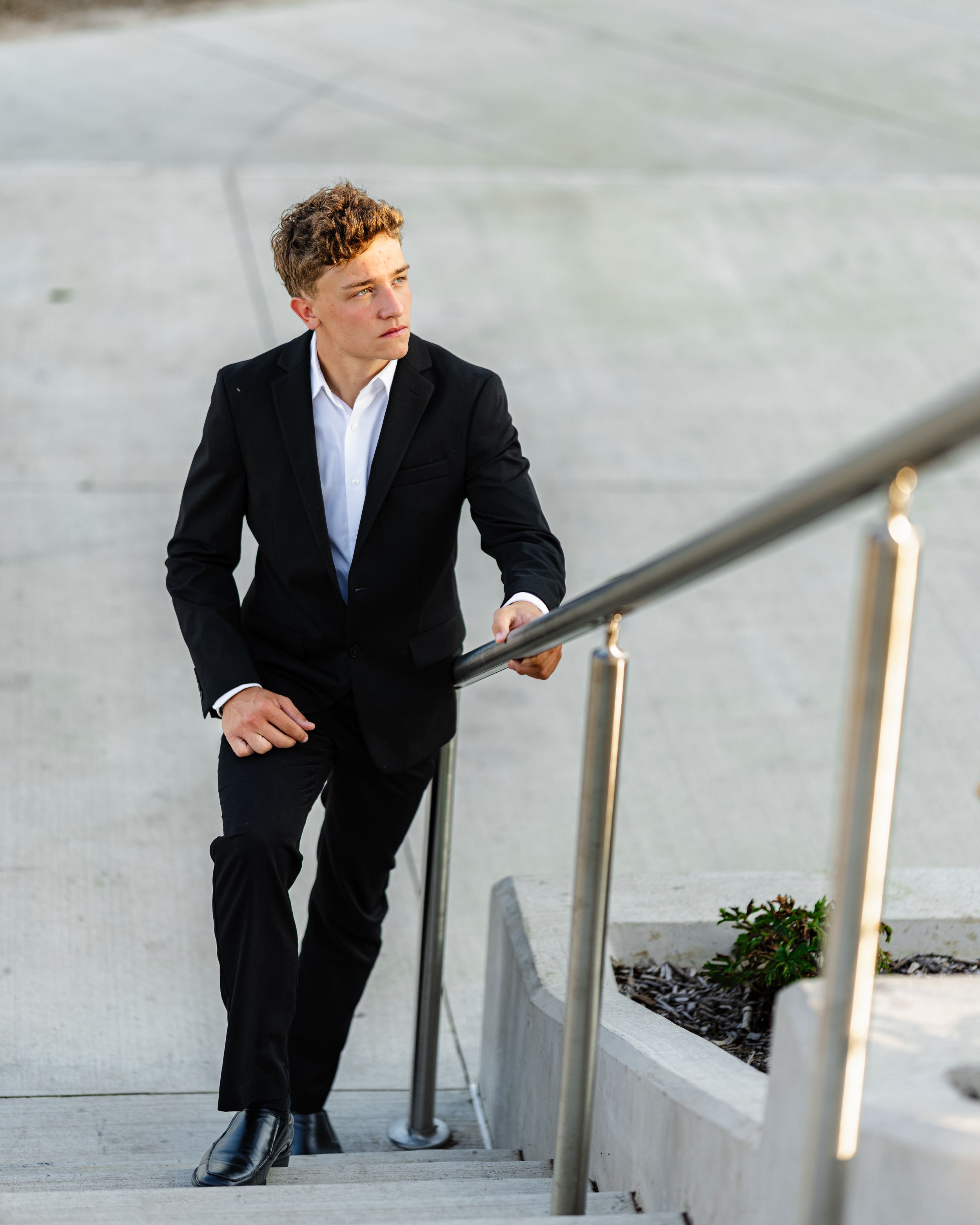 A person in a black suit and white shirt poses on concrete steps with a modern metal railing.