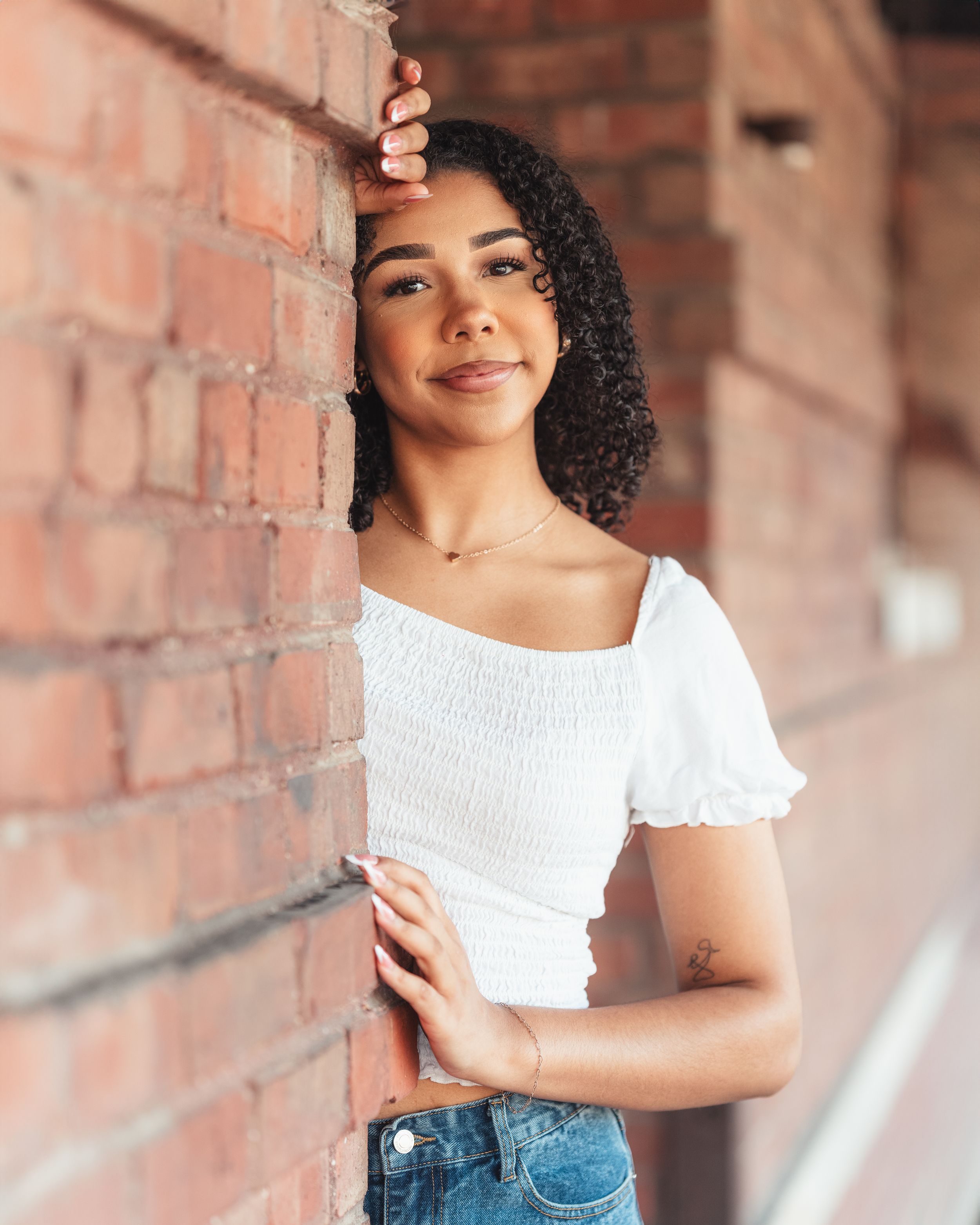 Young person in white top and denim against brick wall smiling in natural outdoor lighting.