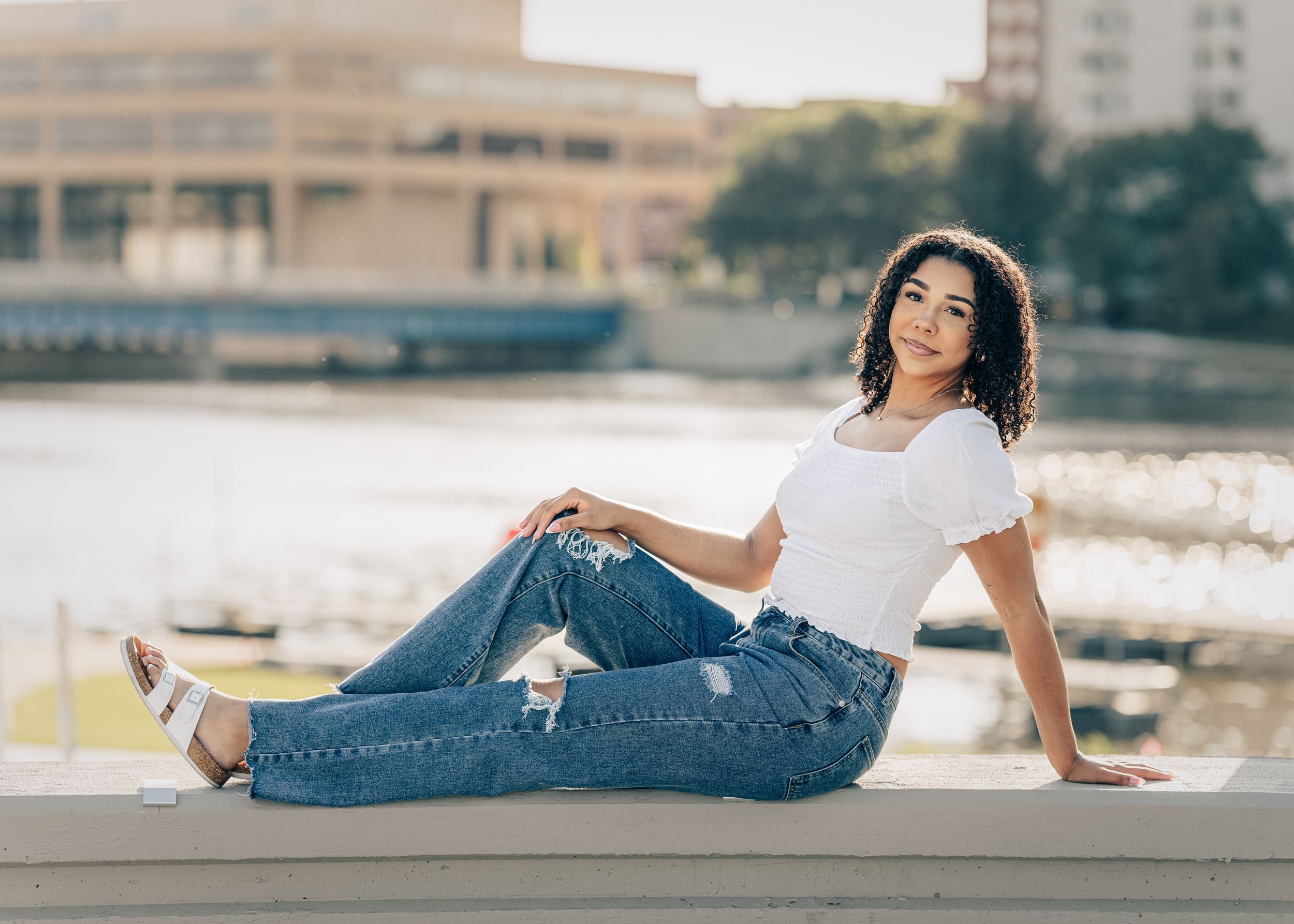 A person in a white t-shirt and distressed jeans relaxes on a concrete ledge near a waterfront with urban buildings behind.