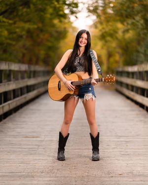Musician playing acoustic guitar while standing on a wooden boardwalk surrounded by autumn trees.