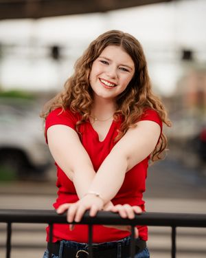 A person in a red top and jeans leans on a railing while smiling outdoors.