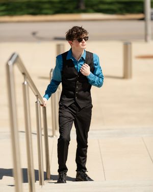 A person in a blue shirt and black vest leans against a railing while holding a phone outdoors.