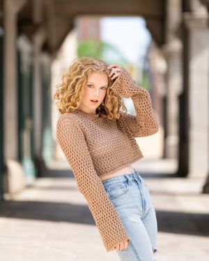 A model in a beige knit sweater and light wash jeans poses against an architectural backdrop.
