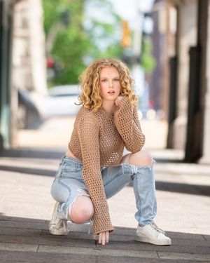 A person in jeans and a brown sweater crouches on a sidewalk in a dynamic urban fashion pose.