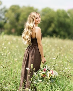 Woman in brown dress stands in wildflower meadow with flowing hair on sunny summer day.