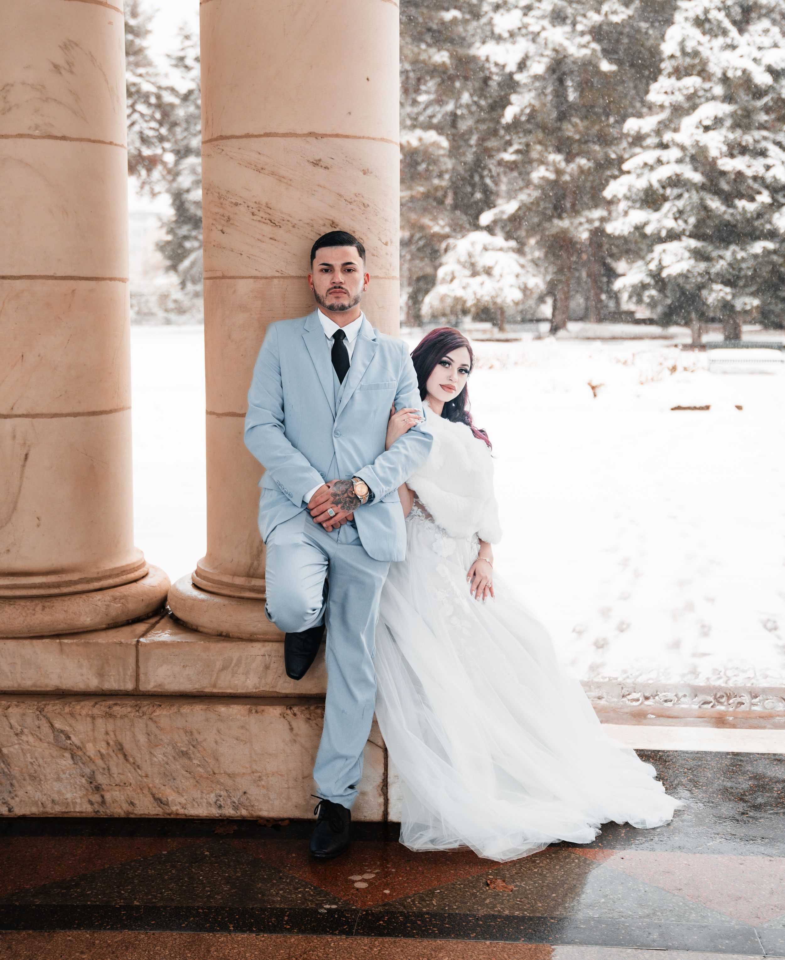 A couple in formal wedding attire sits together on stone steps in a romantic outdoor setting with columns.