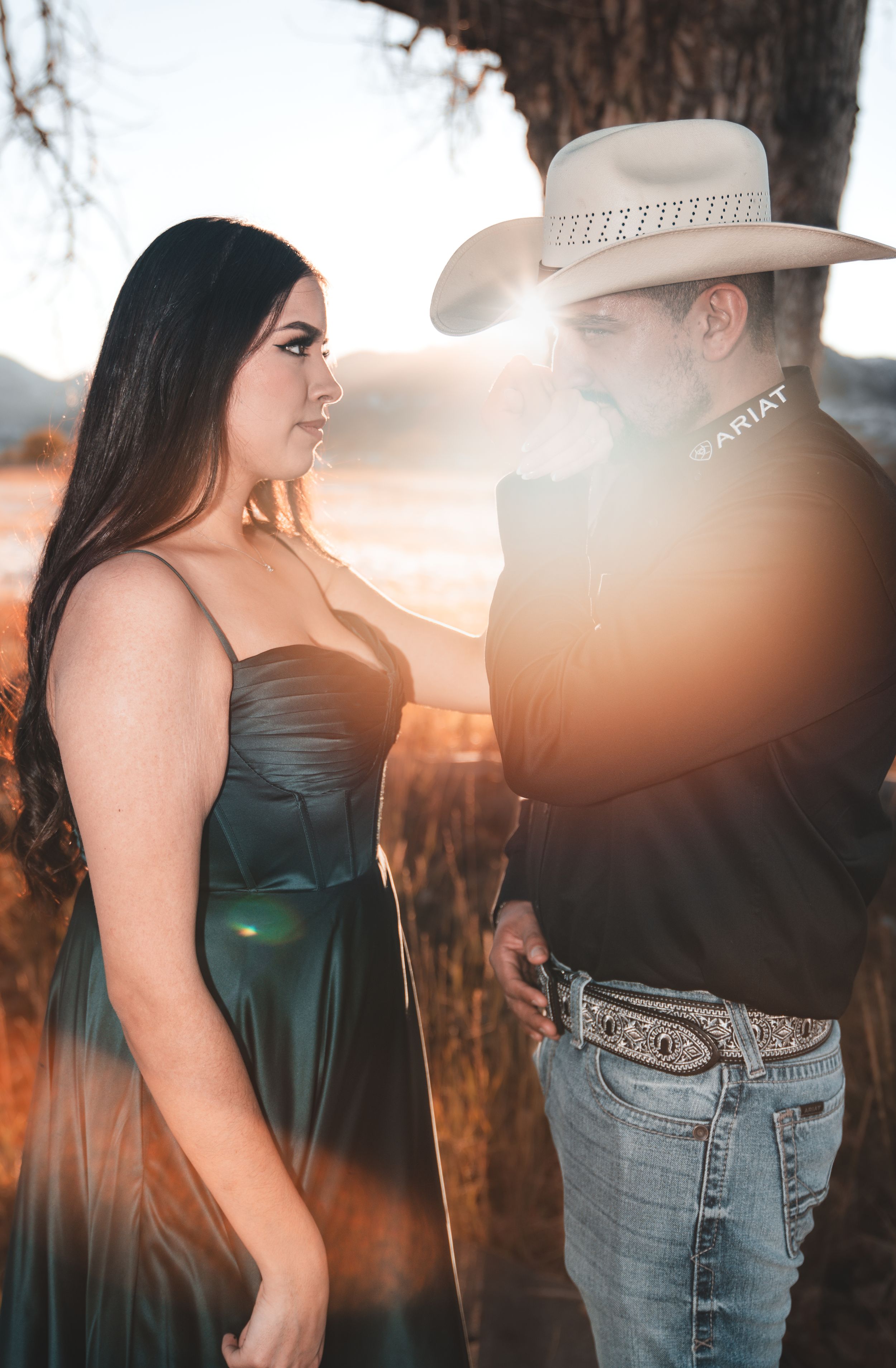 A couple in western attire stands together at sunset, silhouetted against a warm golden sky.