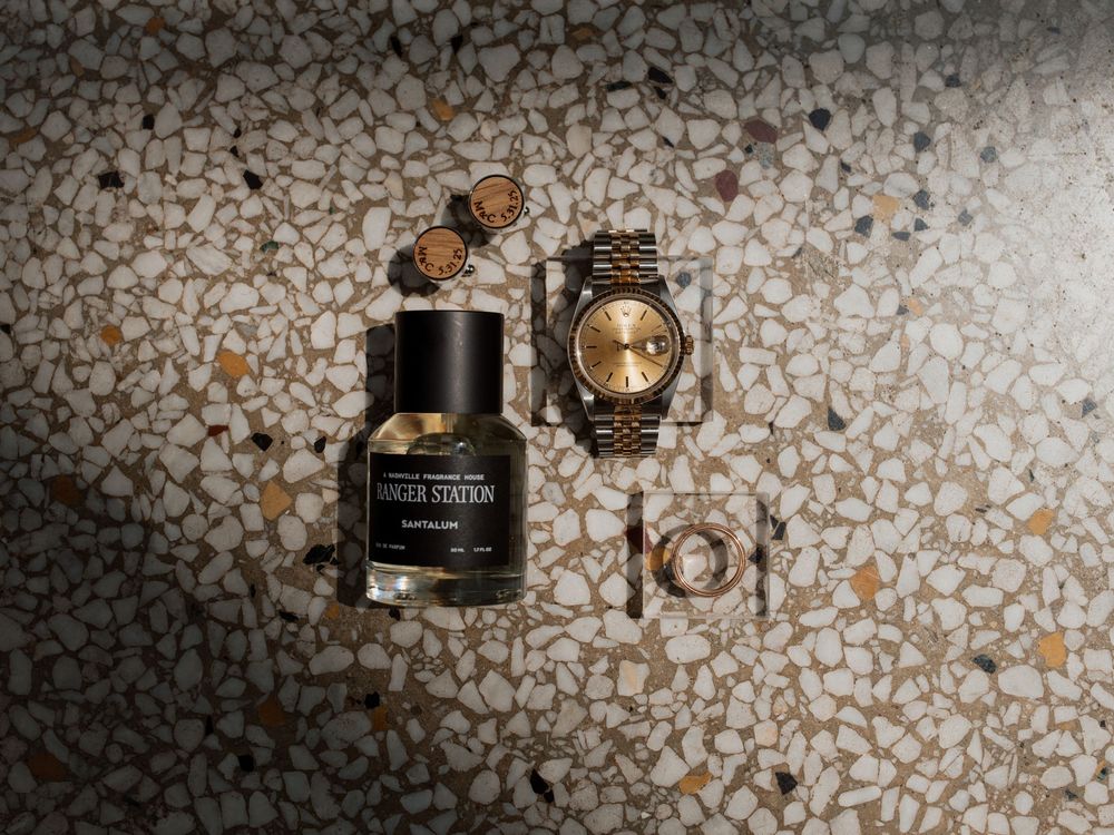 A luxury perfume bottle and gold watch arranged on a textured stone surface with scattered orange petals.
