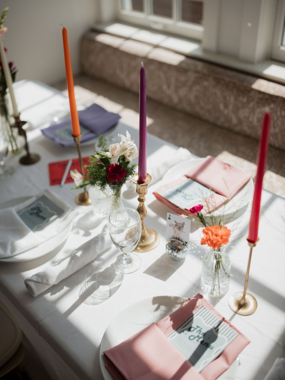 Elegant dinner table setting with pastel pink napkins and colorful taper candles casting shadows on white tablecloth.