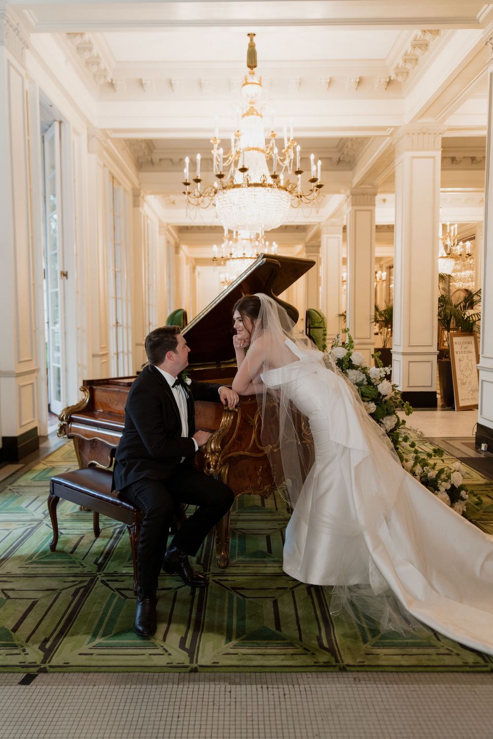 Bride and groom share an intimate moment at a grand piano in an elegant ballroom with crystal chandelier and ornate decor.