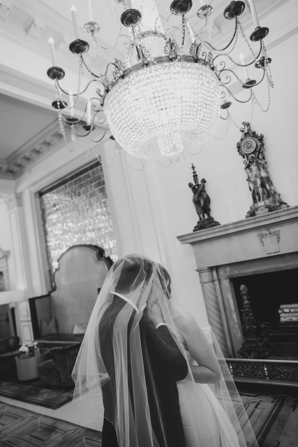 A black and white wedding photography scene with a grand chandelier and ornate fireplace mantle in an elegant room.