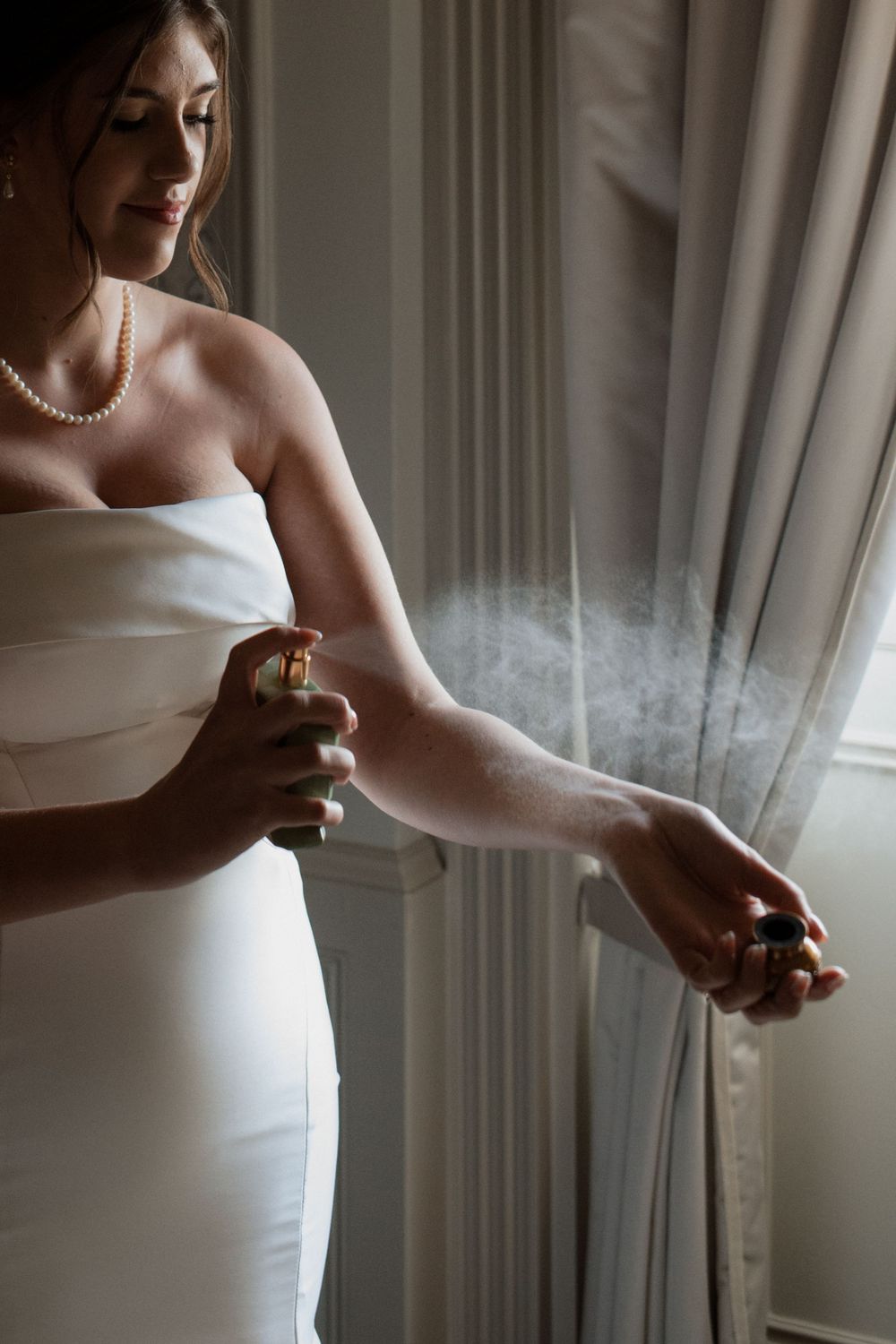 Someone wearing a white wedding dress holds a perfume bottle near a window with sheer curtains in soft lighting.